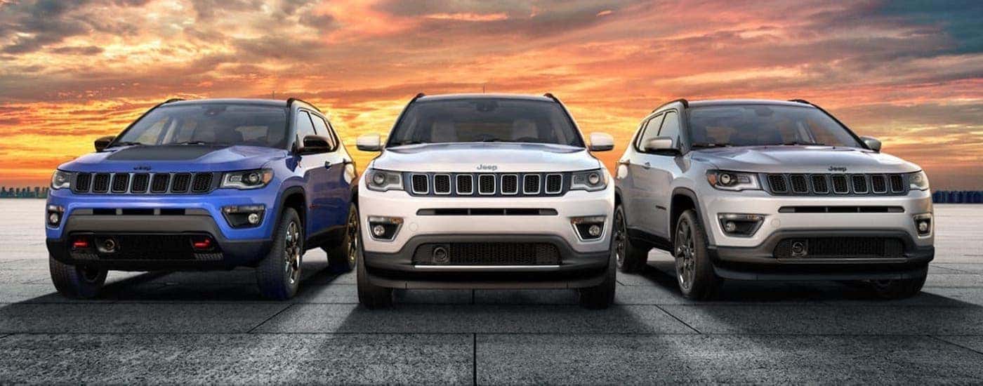 A blue, a white, and a silver 2019 Jeep Compass are parked in front of a vibrant yellow and orange sunset in Colorado Springs, CO.