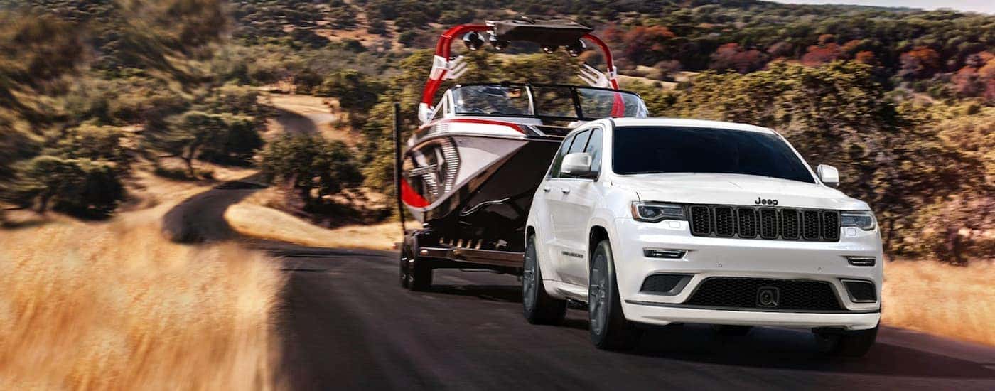 A white 2021 Jeep Grand Cherokee is towing a boat uphill on a rural road.