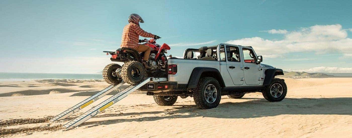 A four wheeler is being driven into the bed of a silver 2021 Jeep Gladiator with no roof that is on…