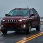 A red 2017 Certified Pre-Owned Jeep Cherokee is driving on a coastal highway in the rain at dusk.