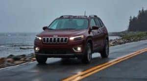 A red 2017 Certified Pre-Owned Jeep Cherokee is driving on a coastal highway in the rain at dusk.