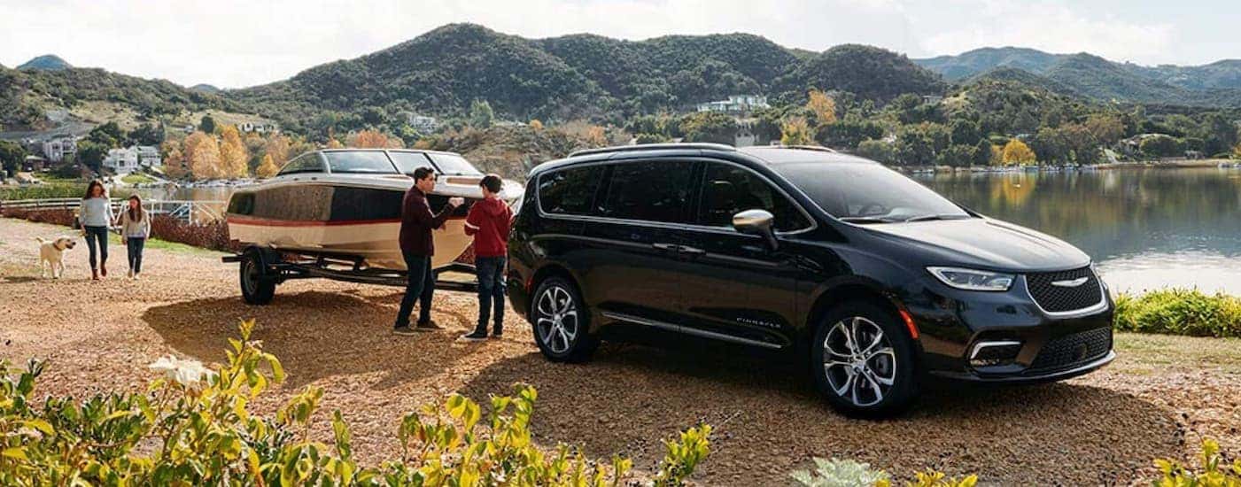 A family is next to a black 2021 Chysler Pacifica with a boat on a dirt road next to a lake.