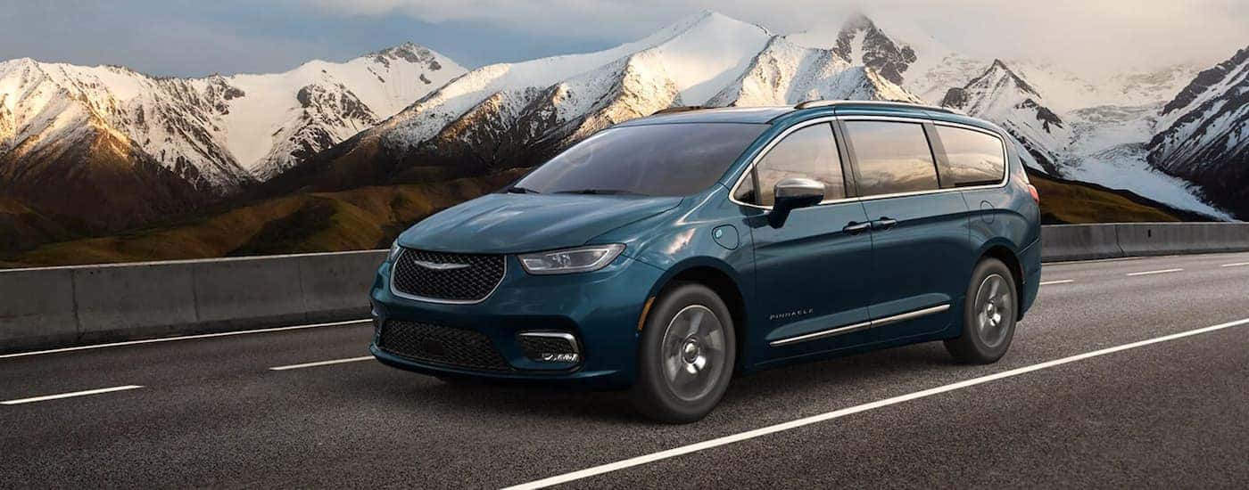 A blue 2021 used Chrysler Pacifica Hybrid is driving on a highway past snowy mountains.