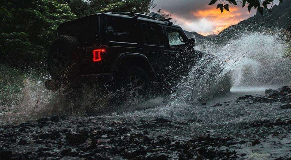 A black Certified Pre-Owned Jeep Wrangler Unlimited splashes through a river at sunset