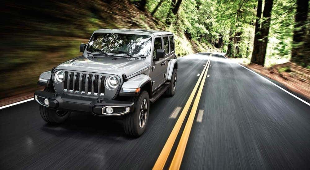 Front-view of a silver 2019 Jeep Wrangler driving fast on a tree lined road.