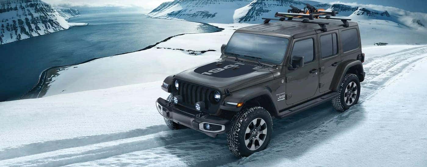 A black 2021 Jeep Wrangler Sahara Unlimited 1941 Edition is shown driving down a snowy trail overlooking mountains and a lake.