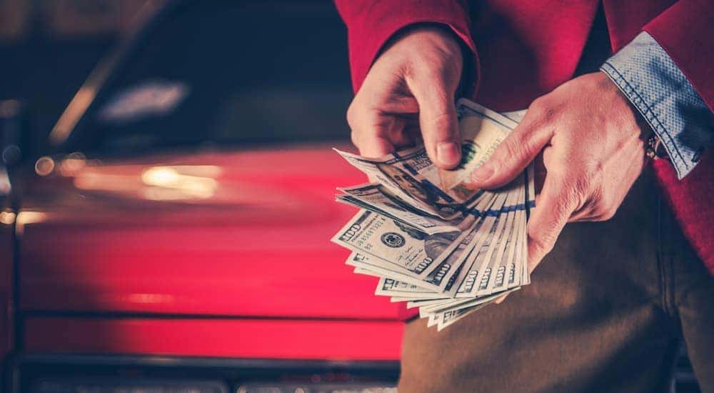 A person is shown holding a handful of cash in front of a red car.