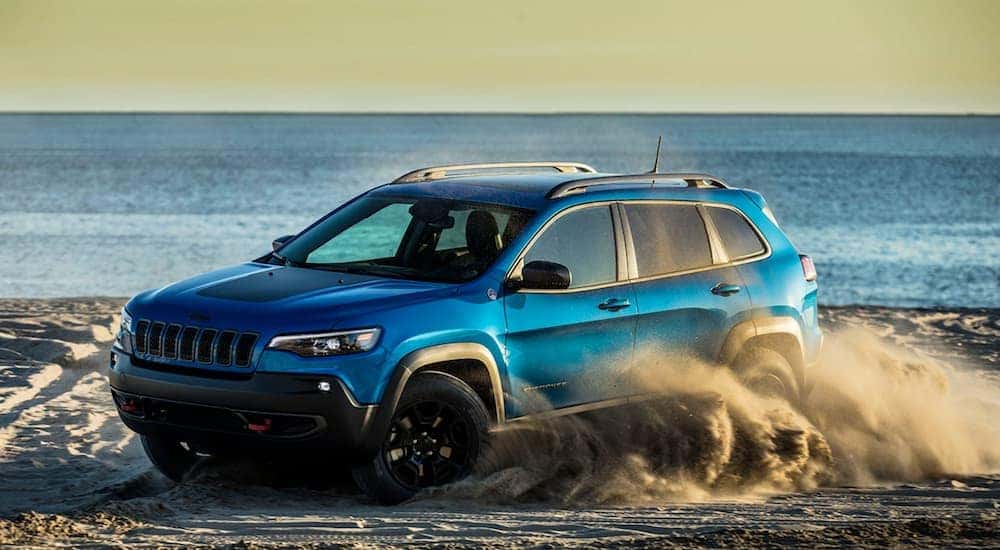 A blue 2020 Jeep Cherokee Trailhawk is shown from the front while kicking up sand on a beach.