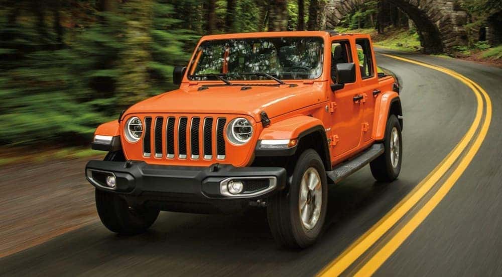 An orange 2020 Jeep Wrangler is shown from the front driving on an open tree lined road.