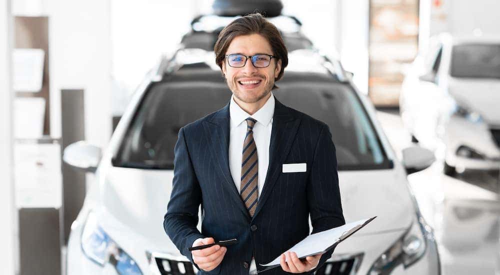 A salesman is shown standing in front of a vehicle while holding paperwork.
