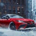 A red 2020 Toyota Camry is shown driving in a snowy city after visiting a dealer with used cars for sale.